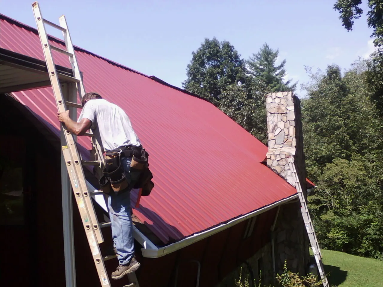 Roofer on a ladder beside a red metal roof and stone chimney — Soffit & Fascia Repair by Roofing Brothers in New Kent